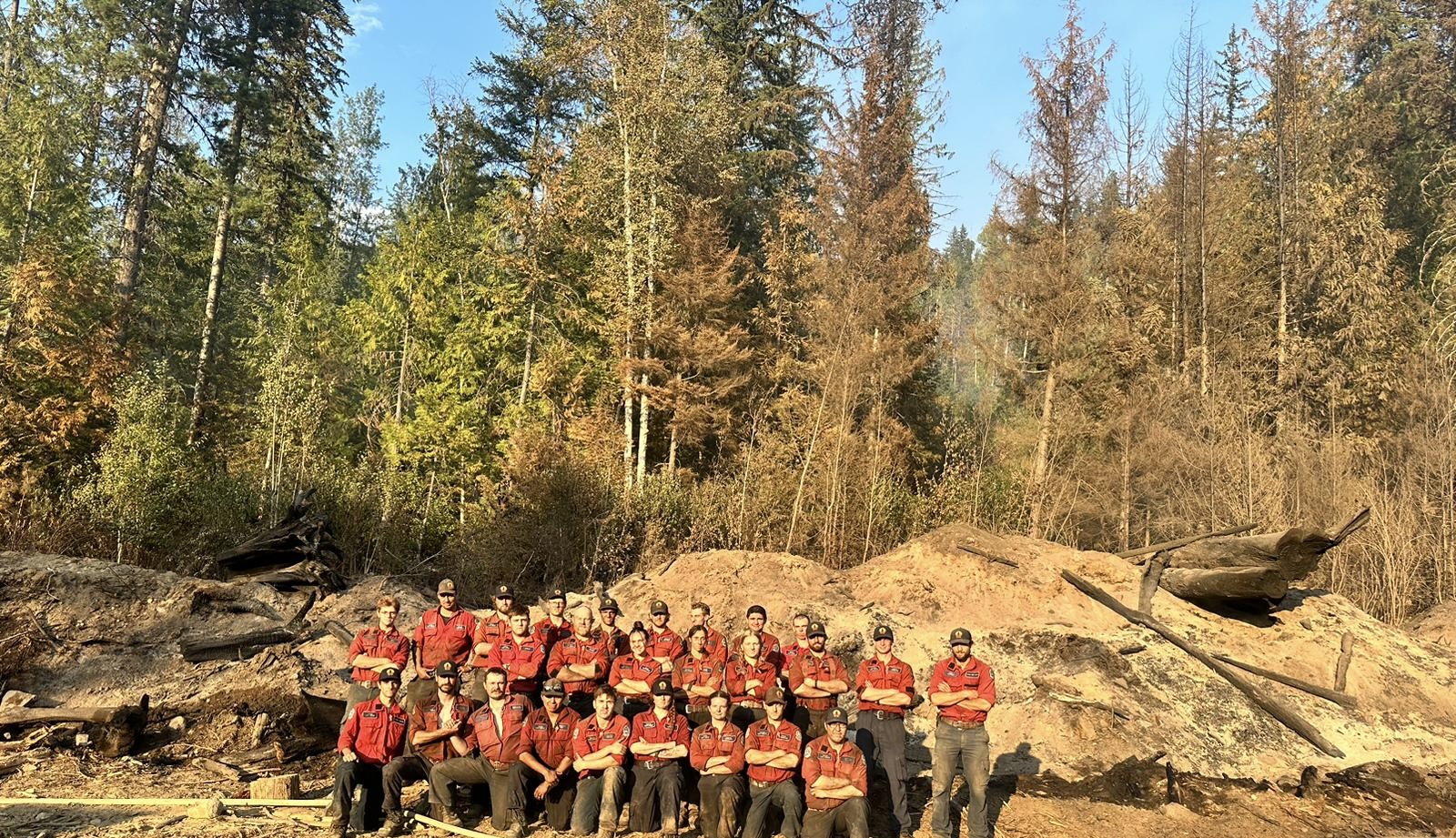 26 people standing in three rows in front of a forest. They are standing in a control area used to help stop wildfires from spreading.