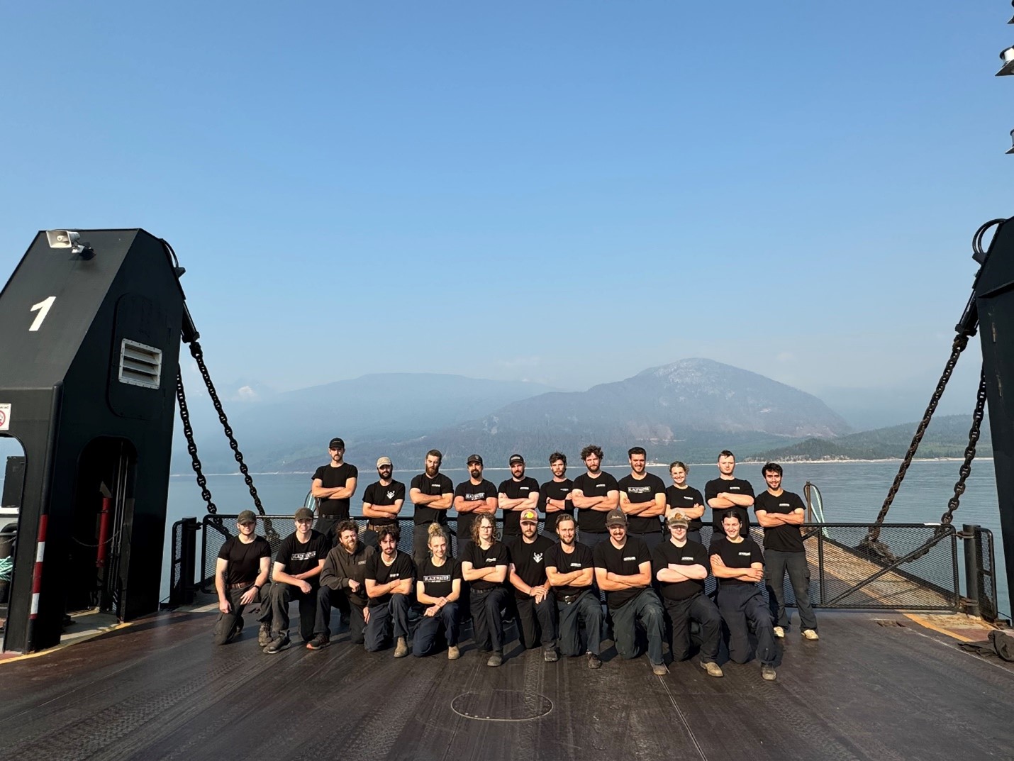 22 people posing in two rows on the end of a boat in a lake. 11 people are standing in the back row with their arms crossed while the other 11 people are kneeling in the front row.