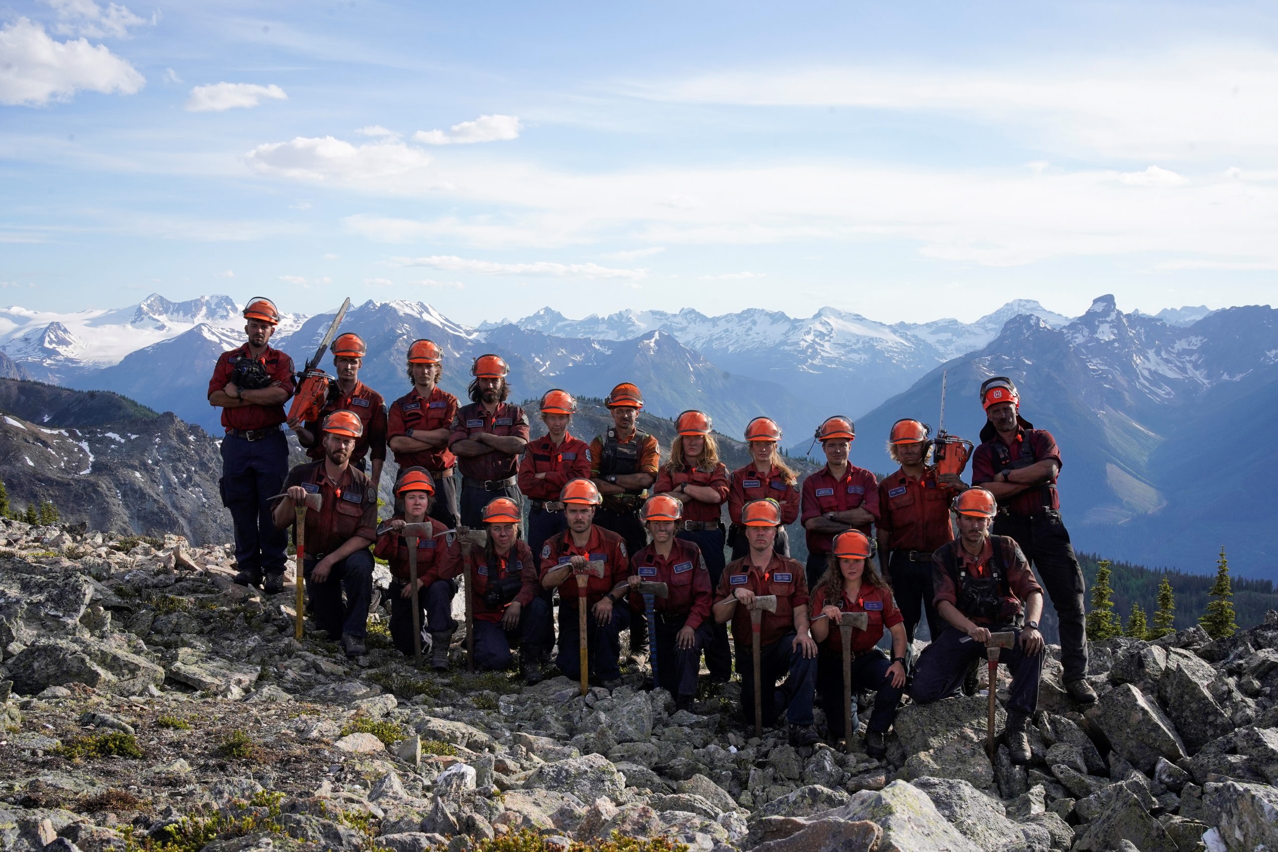 19 people posing on a rocky slope in a valley. 11 people are standing in the back row and eight people are kneeling in the front row. Some people are holding forestry equipment, such as axes or chainsaws, while others just stand with their arms crossed.