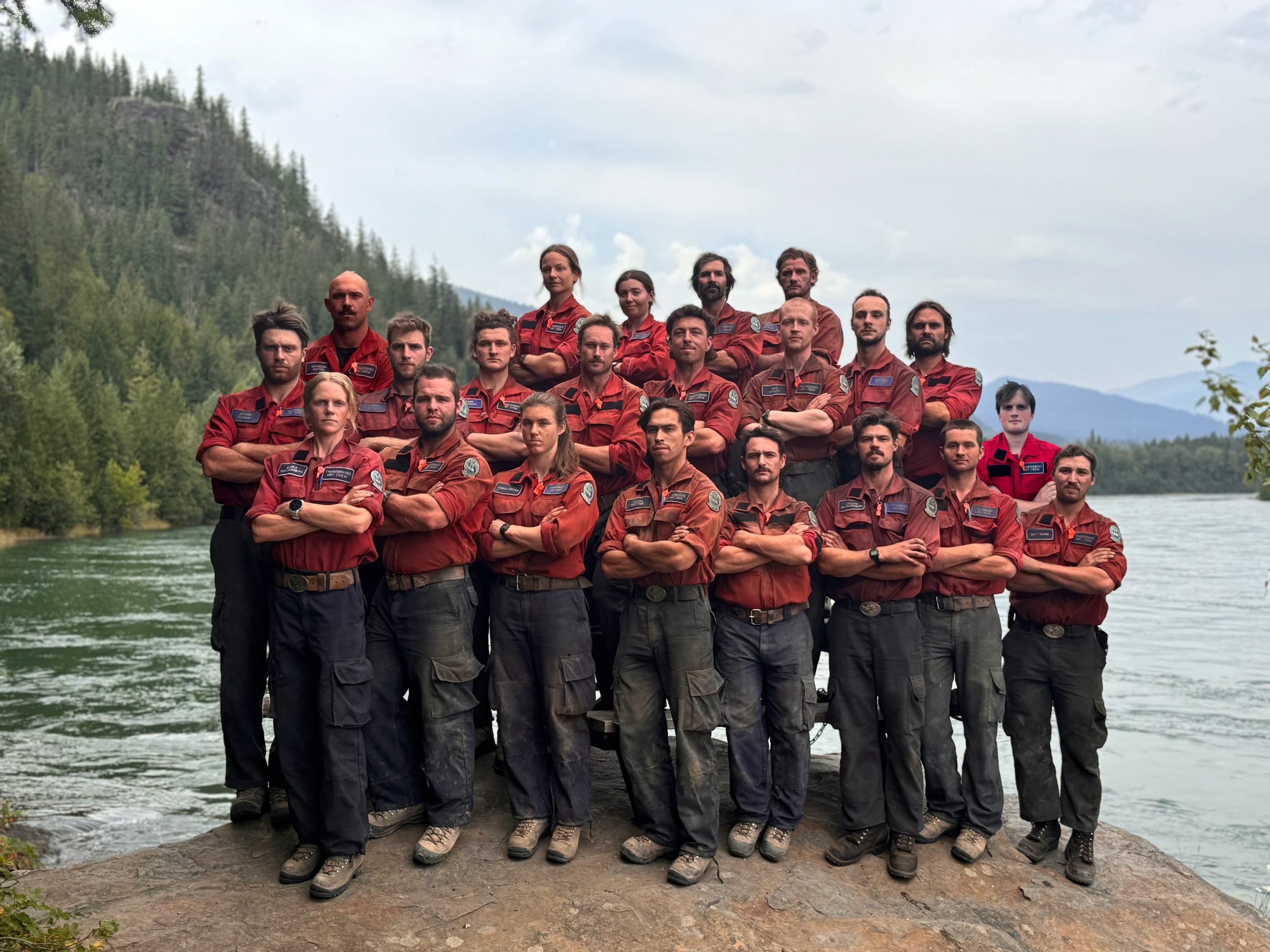 22 people standing in a group with their arms crossed. They are standing on the shore on a lake in a forest.