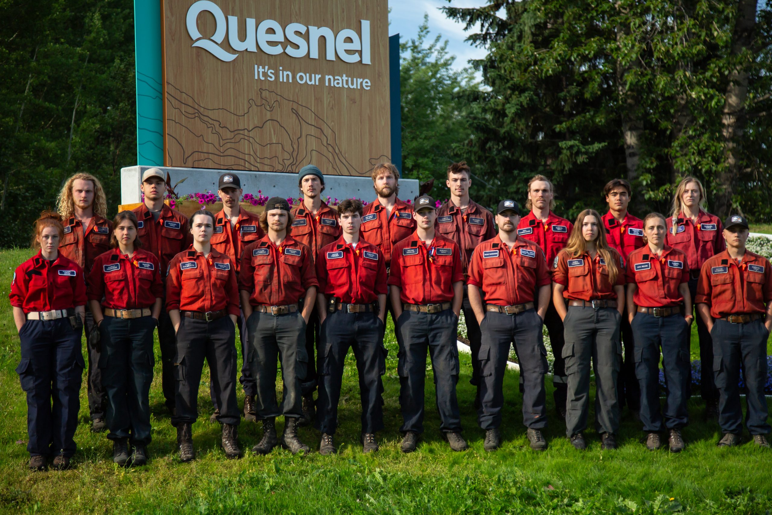 19 people standing in two rows in front of the Quesnel welcome sign. Nine people are in the back row and 10 people are in the front row.