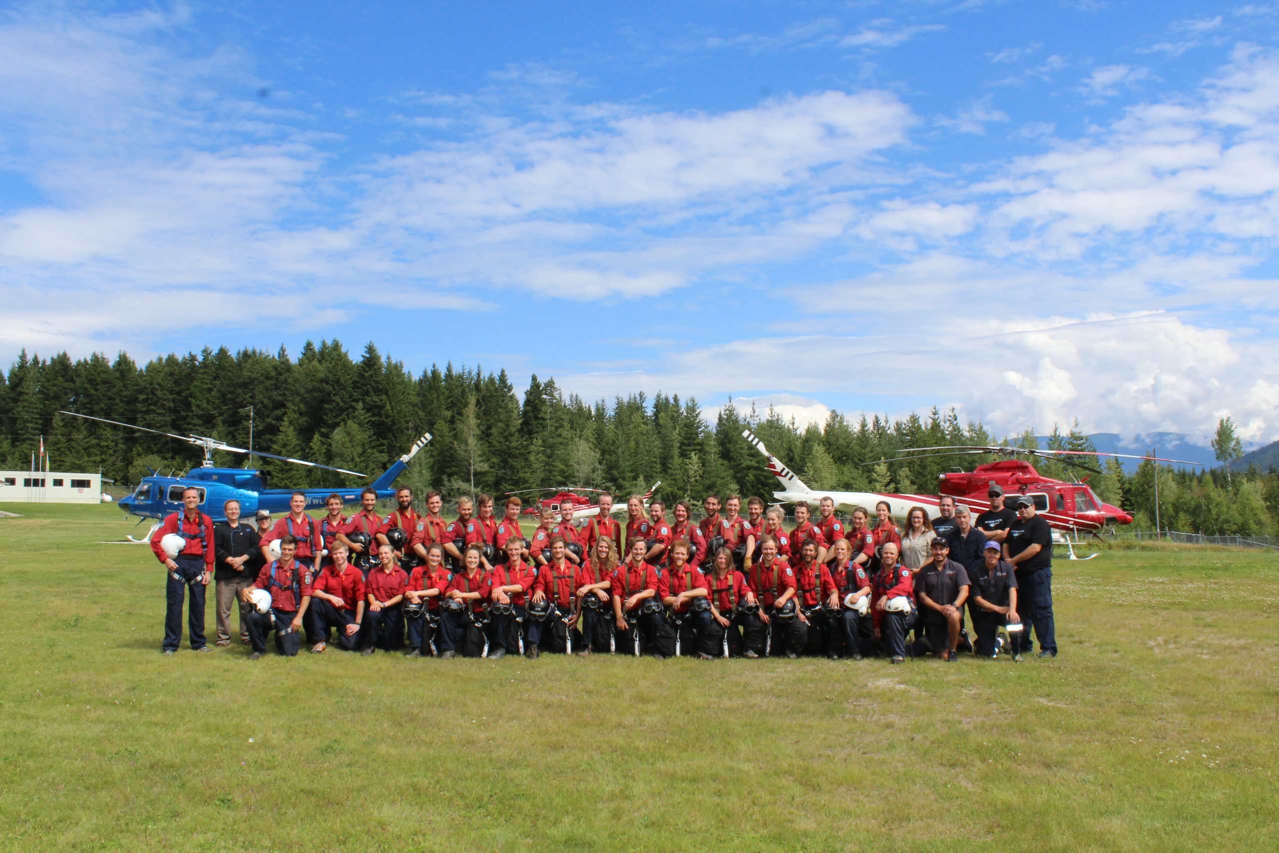 A large group of people posing in 2 rows. They are in a large field next to a forest and there are two helicopters parked behind them.