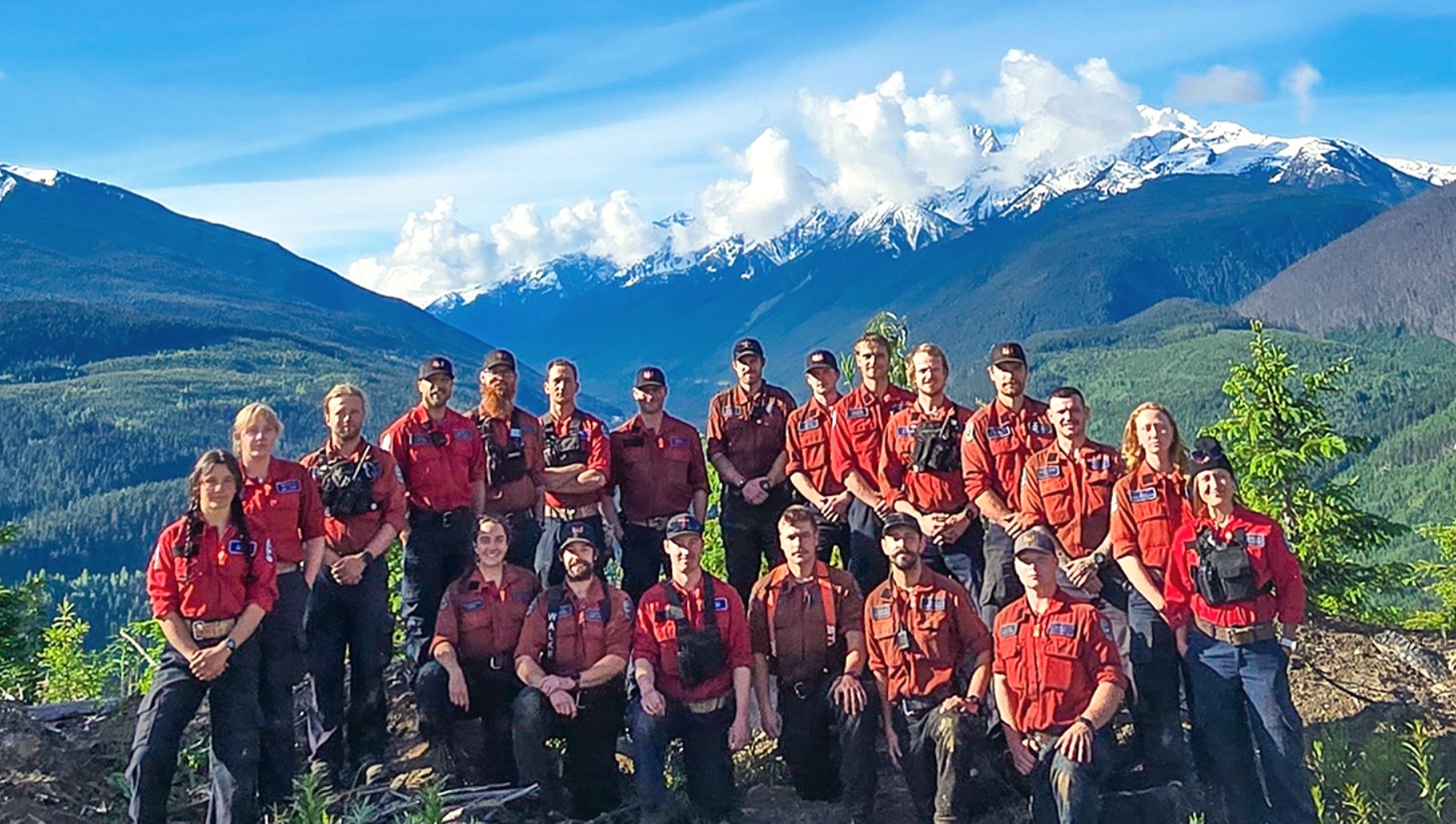 21 people standing on top of a mountain in a forested valley. 15 people are standing in the back row while six people are kneeling in the front row.