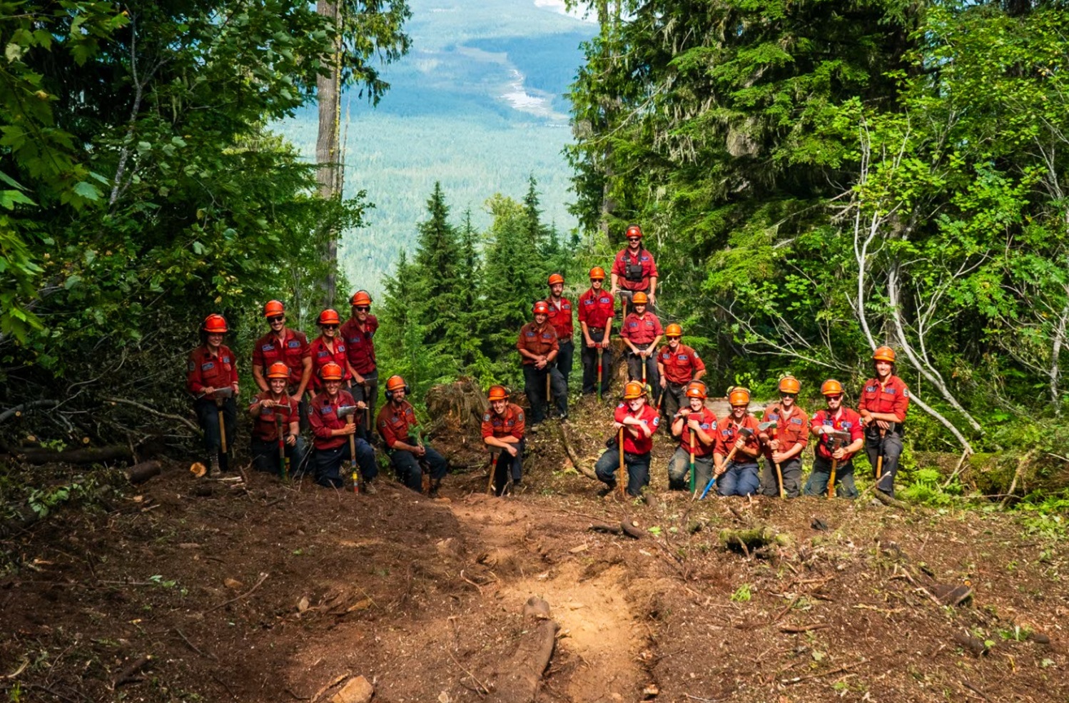 20 people are posing in the middle of a lush forest. Everyone is holding a tool such as an axe or a pickaxe.