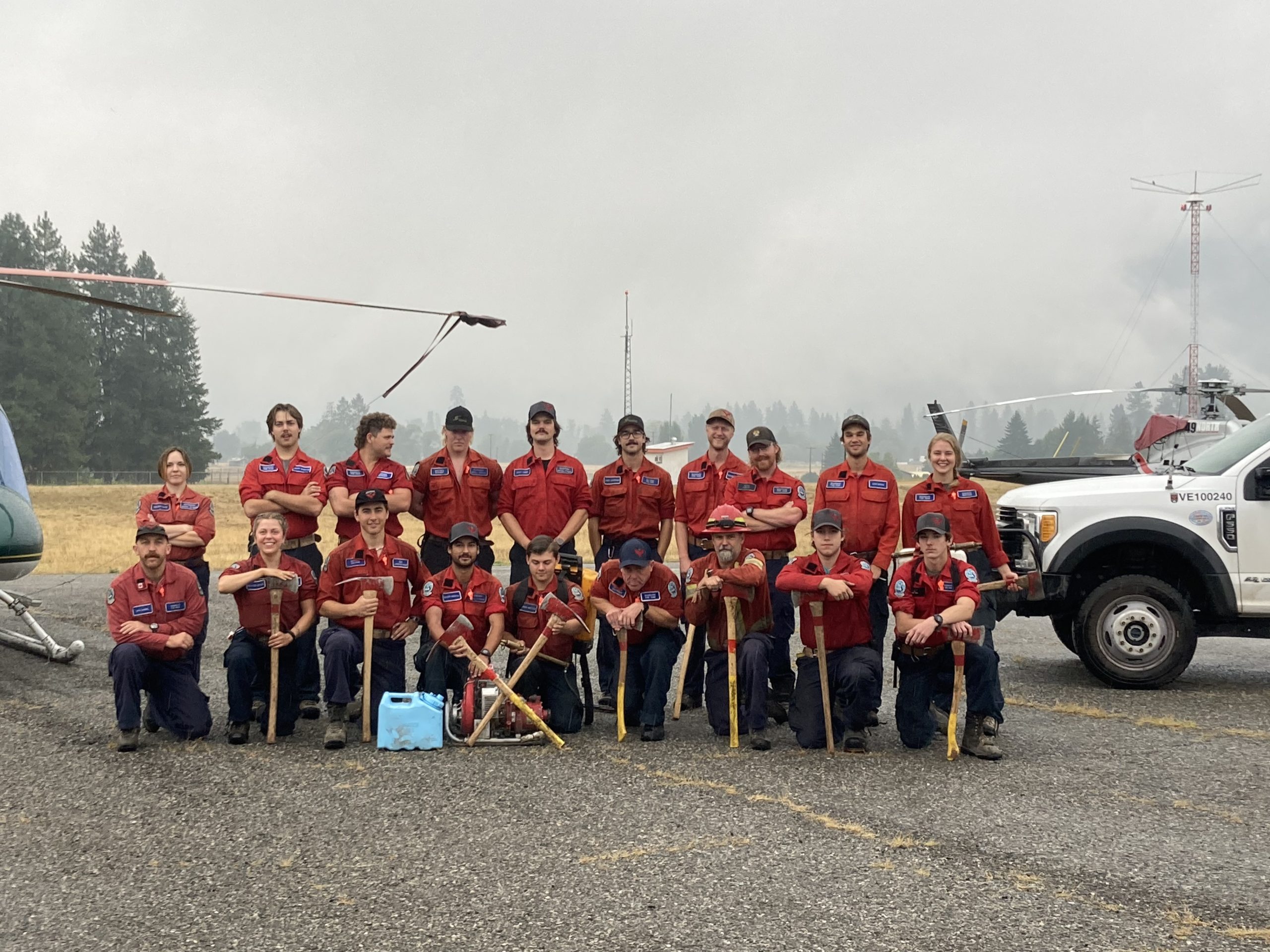 19 people posing on the tarmac of a small airport with a truck and two helicopter seen behind them. 10 people are standing in the back row while the other 9 people kneel in the front row. Eight people in the front row are also holding an axe.