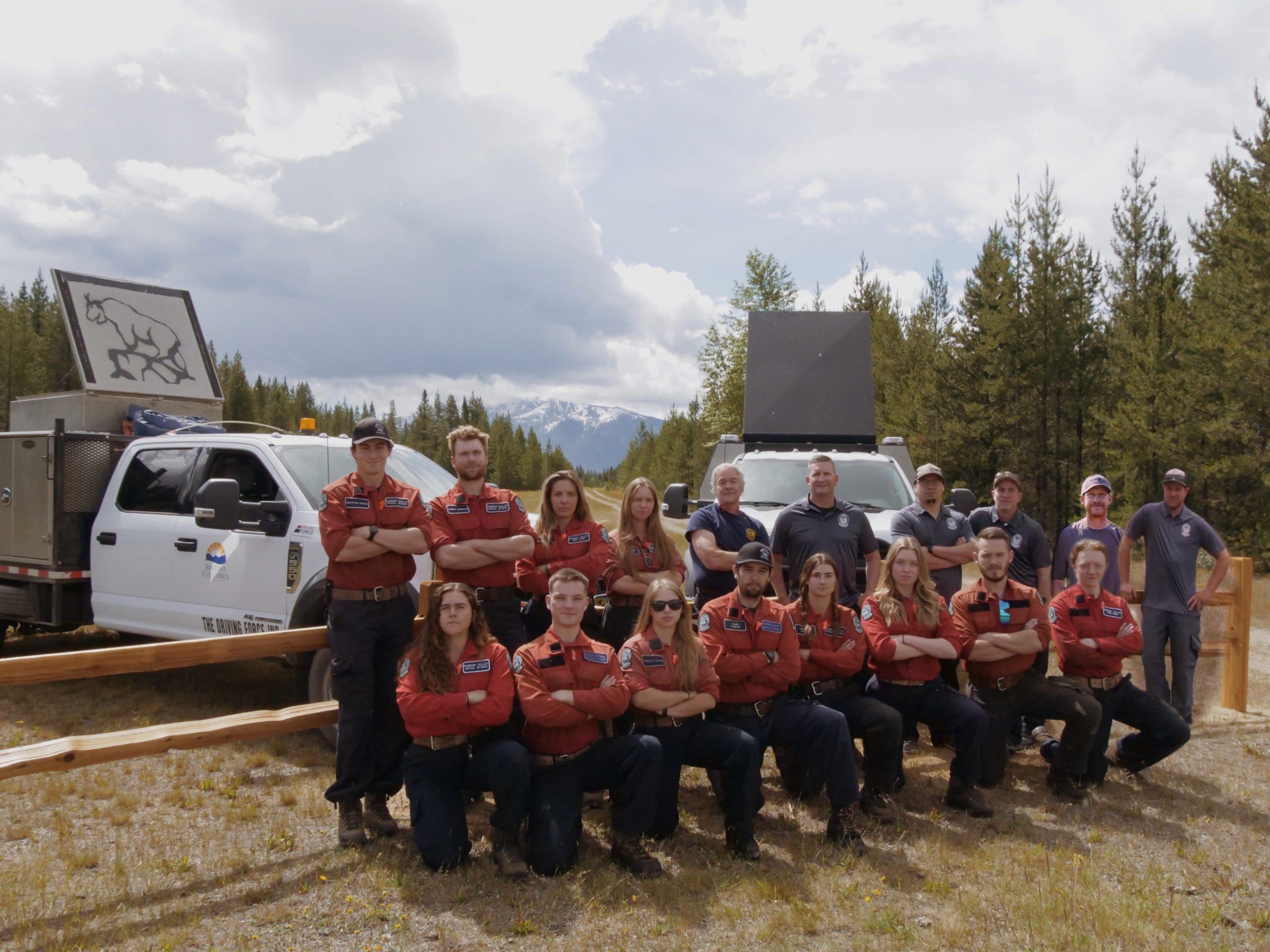 18 people posing in front of a wooden fence in a forest. There are also two trucks behind them with one displaying the Robson Valley IA Crew logo.