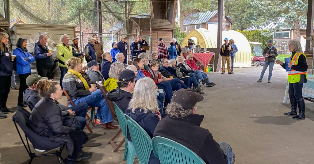 A group of people sitting in chairs listening to and looking at a person speaking at the front of the room.