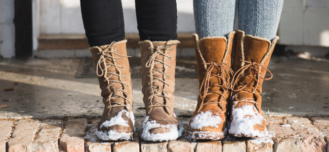 Two people standing on a brick porch. The photo just shows their boots which have the toes covered in snow.