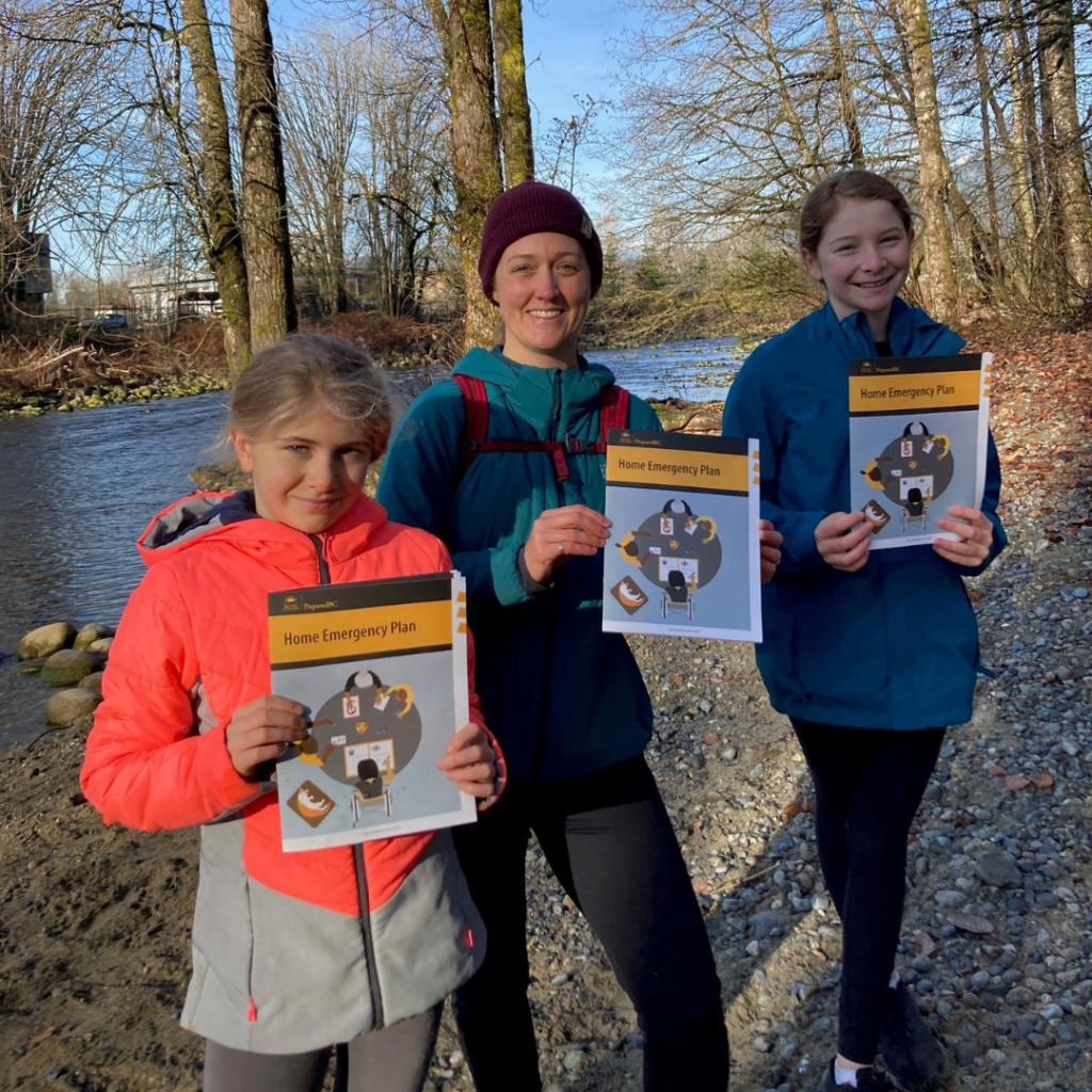 Three people standing along a river bank holding copies of the PreparedBC Home Emergency Plan.