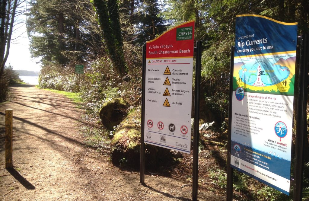 Two large signs with information about coastal hazards are standing next to a trail leading to the coast.